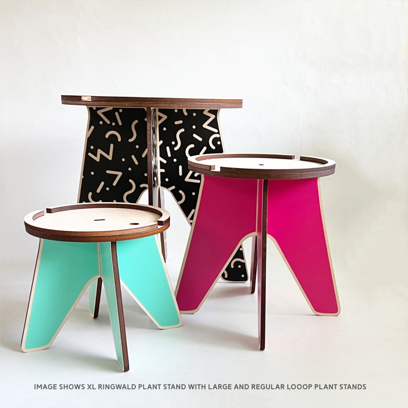 Op de afbeelding: Drie plantenstandaards in verschillende maten. De grootste standaard is zwart met witte geometrische patronen. De middelste standaard is roze en de kleinste standaard is turquoise.  "IMAGE SHOWS XL RINGWALD PLANT STAND WITH LARGE AND REGULAR LOOP PLANT STANDS"