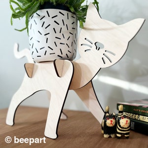 May include: A wooden cat planter stand with a white planter featuring black dashes. The stand is holding a small green plant. Two small wooden cat figurines are in the foreground.