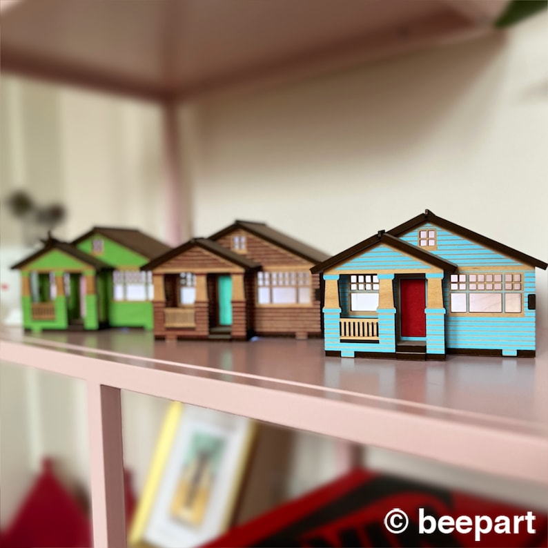 May include: Four miniature wooden houses in different colors, including blue, green, brown, and beige, are arranged on a shelf. The houses have small windows and doors, and some have porches.