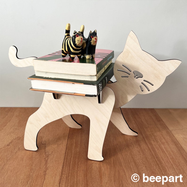 May include: A wooden cat figurine bookend with a whimsical design. The cat is carrying a stack of books and has a pair of small, colourful wooden cats on top.