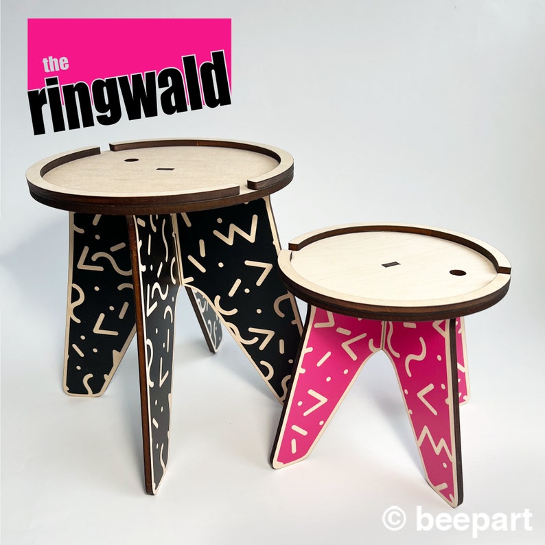 May include: Two wooden stools with geometric patterns. The larger stool has a black and white pattern and the smaller stool has a pink and white pattern. The text "the ringwald" is printed in pink on a white background.