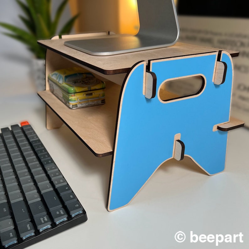 May include: A light blue wooden laptop stand with a shelf for storage. The stand has a cutout handle and is made of plywood. The stand is sitting on a white surface.