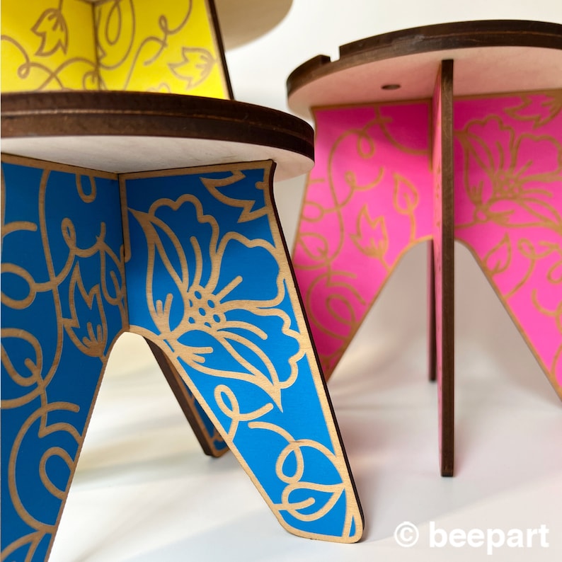 May include: Two small wooden stools with a floral design. The stool on the left has a blue base with gold floral accents. The stool on the right has a pink base with gold floral accents.
