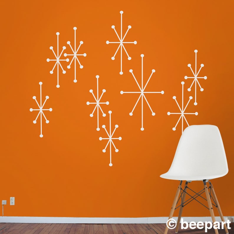 Puede incluir: Un conjunto de nueve pegatinas de pared blancas en forma de estrella sobre una pared naranja. Las pegatinas est&aacute;n dispuestas en un patr&oacute;n aleatorio. Una silla blanca est&aacute; en primer plano.