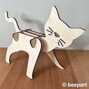 May include: A wooden cat-shaped desk organiser with three slots for pens or pencils. The cat is light brown with black details and a cute face.