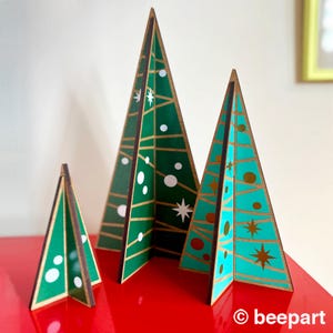 Peut inclure: Trois sapins de Noël en bois de différentes tailles, décorés de motifs géométriques en vert, turquoise, or et blanc. Les sapins sont posés sur une surface rouge.