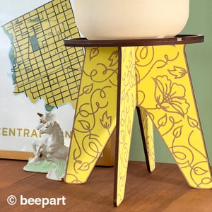 May include: A yellow wooden plant stand with a floral pattern. The stand has three legs and a round top.