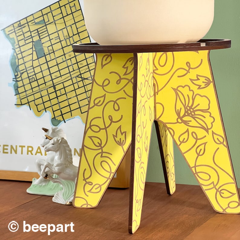 May include: A yellow wooden plant stand with a floral pattern. The stand has three legs and a round top.
