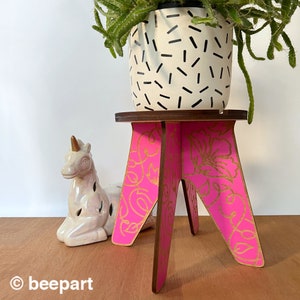 May include: A pink wooden plant stand with gold floral designs and three legs. A white ceramic unicorn figurine is in the foreground. A potted plant with black and white patterned pot is on top of the stand.