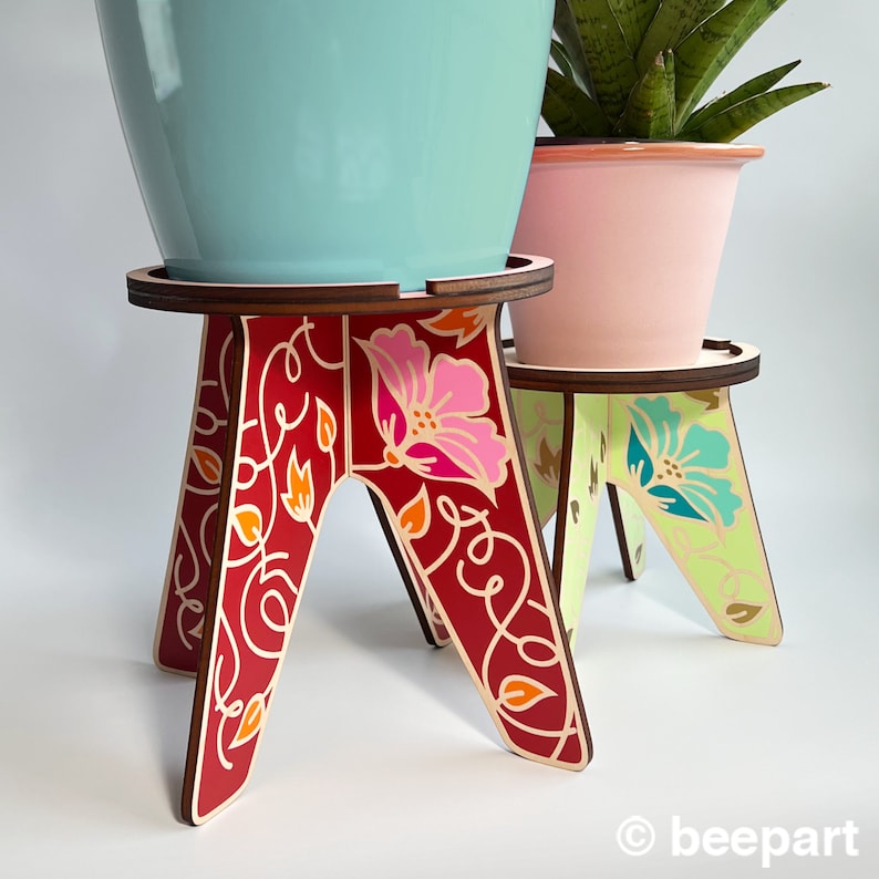 May include: Two wooden plant stands with floral designs. The larger stand is red with pink flowers and the smaller stand is green with blue flowers. Both stands have a light wood top.