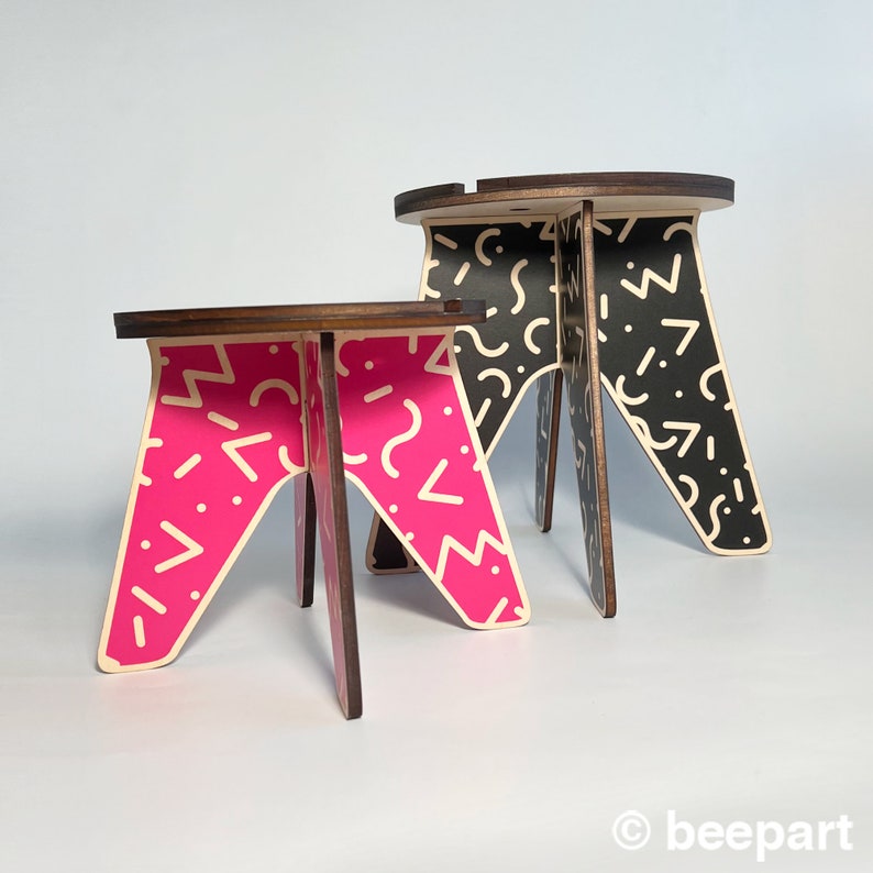 May include: Two wooden stools with geometric patterns. The larger stool is black with white geometric shapes. The smaller stool is pink with white geometric shapes.