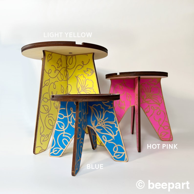 May include: Three wooden stools with floral designs. The largest stool is light yellow with gold accents, the middle stool is blue with gold accents, and the smallest stool is hot pink with gold accents.