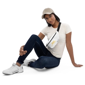 Könnte beinhalten: Eine weiße Bauchtasche mit gelbem "GOOD VIBES"-Text und einem schwarzen verstellbaren Riemen. Die Bauchtasche wird von einer Person getragen, die ein weißes T-Shirt, blaue Jeans und eine beige Baseballkappe trägt.