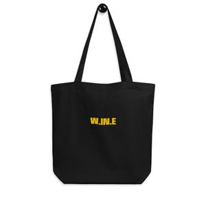 May include: Black tote bag with the word "W.IN.E" printed in yellow. The bag has two black handles and is hanging from a black hook. The bag is made of a soft material.