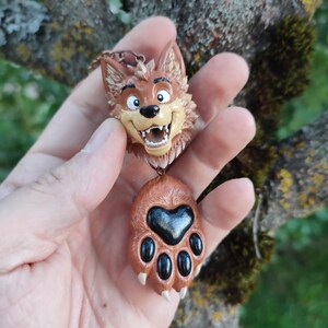 Könnte beinhalten: Ein brauner Wolf-Anhänger bestehend aus einem modellierten Wolfskopf mit lächelndem Ausdruck und offenem Maul, verbunden durch eine kupferfarbene Kette mit einem passenden braunen Pfotenabdruck mit großem schwarzem Herzballen und vier schwarzen Zehen.
