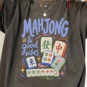 Könnte beinhalten: Ein anthrazitfarbenes Kurzarm-T-Shirt mit einem Grafikdruck, der den Text "MAHJONG & good vibes" in Blau zeigt. Das Design enthält verschiedene bunte Mahjong-Steine mit chinesischen Schriftzeichen und Blumenmustern sowie kleine Sternsymbole.