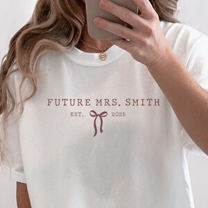 Puede incluir: Camiseta blanca de gran tamaño con el texto "FUTURE MRS. SMITH EST. 2023" y un gráfico de lazo rosa.