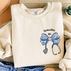 Puede incluir: Una sudadera de color crema con un diseño de lazo azul y blanco y un estetoscopio. El texto "Samantha RN" está impreso en la sudadera.