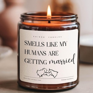 Puede incluir: Una vela de vidrio marrón con una etiqueta blanca que dice "Smells Like My Humans Are Getting Married". La vela está encendida y tiene una ilustración en blanco y negro de dos corazones entrelazados en la etiqueta. La etiqueta también incluye el texto "KAIROA CANDLES", "Burn time: 50 hours oz" y "Midnight blackberry".