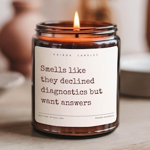 May include: A brown glass candle with a white label that reads "Smells like they declined diagnostics but want answers". The candle is lit and has a burn time of 20 hours. The scent is Midnight Blackberry.