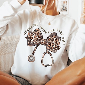 Puede incluir: Sudadera blanca con un lazo de estampado de leopardo y un estetoscopio, con el texto "Neonatal Intensive Care Unit".