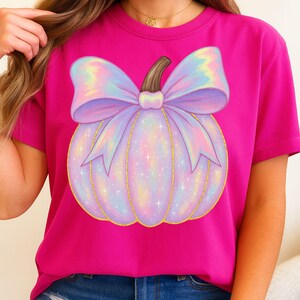 May include: A magenta t-shirt featuring a whimsical pumpkin design. The pumpkin is a soft lavender color with a large, iridescent bow and gold glitter accents. The stem is brown.