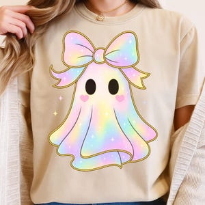 Może przedstawiać: Beżowa koszulka z nadrukiem kreskówki ducha. Duch ma dużą pastelową tęczową kokardę, czarne oczy i różowe policzki w kształcie serca. Małe różowe gwiazdki są rozrzucone.