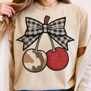 May include: Beige t-shirt featuring an embroidered cherry design. The design includes a black and white checkered bow, a brown and tan patterned cherry, and a red cherry. The shirt has a crew neck and short sleeves.