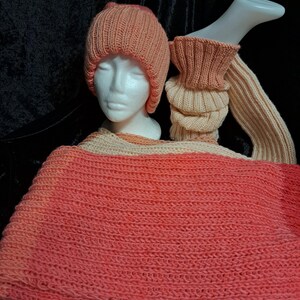 May include: A knitted peach-coloured hat, scarf, and arm warmers set. The hat is ribbed, and the scarf and arm warmers have a textured design. The set is displayed on a mannequin head and arm, against a dark background.