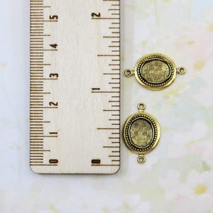 May include: Two antique gold-colored oval connector charms with a woven pattern center and a dotted border. Each charm has two small loops for attaching to other components. A wooden ruler with inch markings is in the background.