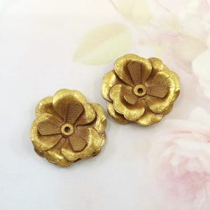 Op de afbeelding: Twee goudkleurige bloemvormige versieringen. Elke bloem heeft meerdere lagen bloemblaadjes en een gestructureerd midden met een klein gaatje. De bloemen zijn gemaakt van een metalen materiaal, waarschijnlijk voor knutselen of decoratie.