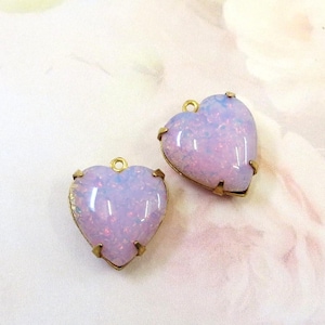 May include: Two heart-shaped pendants with a shimmering, opalescent appearance in shades of lavender and pink. Each heart is encased in a gold-colored metal setting with a small loop at the top for hanging.