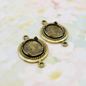 May include: Two antique brass-colored pendant trays with a woven pattern inside the bezel. Each pendant has a small loop at the top and bottom for attaching to a chain or other jewelry component. The bezels are surrounded by a beaded border.