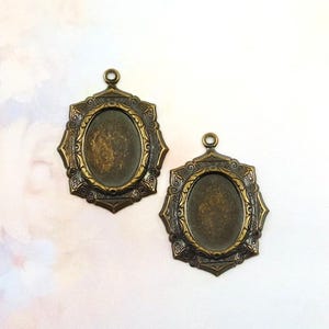 Pode incluir: Duas bases de pingente ovais em tom bronze antigo com bordas ornamentadas em filigrana. Cada base tem um pequeno laço na parte superior para prender a uma corrente ou outro componente de joalheria. As bases estão vazias, prontas para um cabochão ou outra inserção.