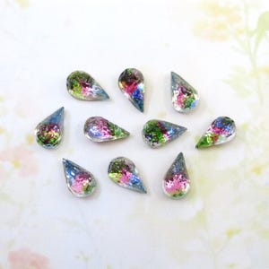 Może przedstawiać: Kolekcja dziesięciu iryzujących szklanych koralików w kształcie łezki. Każdy koralik ma mieszankę kolorów, w tym różowy, zielony i niebieski, z przezroczystą, odblaskową powierzchnią. Koraliki są rozrzucone na białej powierzchni.