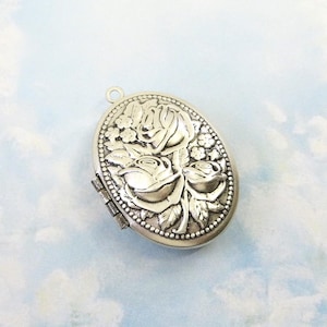 May include: Silver-toned oval locket with a floral rose design. The locket features three roses in the center, surrounded by leaves and small flowers. The border is detailed with small beads, and it has a small loop at the top for a chain.