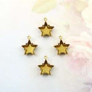 May include: Four gold-colored star charms with a loop at the top for hanging. The stars have a textured surface and are arranged on a white and pink floral background. These charms are suitable for jewelry making or craft projects.