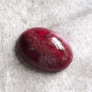 May include: A polished, oval-shaped gemstone with a deep red hue, speckled with gold and lighter red inclusions. The surface is glossy, reflecting light. The gemstone is set against a neutral gray background.