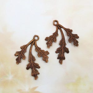 May include: Two copper-toned metal charms, each shaped like a small branch with three detailed oak leaves. Each charm features a small circular loop at the top for attachment, with a textured, antiqued finish highlighting the leaf veins.