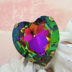May include: A large, heart-shaped iridescent crystal with a vibrant display of colors, including green, purple, and orange. The crystal is faceted, catching light and creating a dazzling effect. Small gold-colored prongs secure the crystal.
