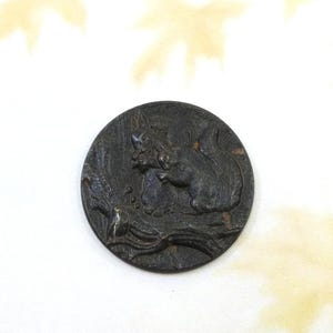 May include: A dark, circular button with a detailed relief of a squirrel, a bird, and foliage. The button is a dark, metallic color, and the design is raised, creating a textured surface. The button is likely made of metal.