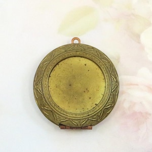 May include: A vintage, round brass locket with an ornate, engraved border. The locket has a hinged opening and a small loop at the top for a chain. The interior is a tarnished gold color. A classic piece of jewelry.