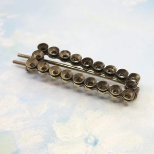 May include: A silver-coloured barrette with a rectangular shape. It has a chain-like design made of small, cup-shaped elements. Two parallel pins extend from one end for securing the barrette in the hair.