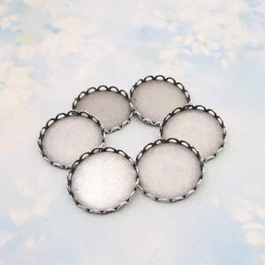 May include: A set of six circular silver-toned metal cabochon settings. Each piece features a smooth, flat, light-colored interior base surrounded by a delicate, scalloped, or crown-like edge with small, evenly spaced cutouts along the rim.