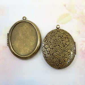 May include: Two antique bronze oval lockets. One locket is open, revealing an empty interior. The other locket is closed, with a floral pattern on the exterior. Both lockets have a small loop at the top for a chain.