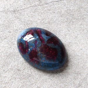 May include: An oval-shaped, glossy stone with a swirling pattern of blue, burgundy, and gold flecks. The stone has a smooth, polished surface and reflects light, creating a shimmering effect. The stone is set against a neutral gray background.