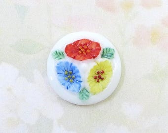 May include: A white, round decorative button with three colorful flowers. The flowers are red, blue, and yellow, with green leaves. The button is likely made of ceramic or glass.