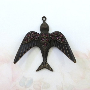 May include: A bronze-toned bird pendant with outstretched wings, embellished with small, round, red gemstones. The bird features a loop for hanging and detailed wing feathers. Suitable for jewellery making or crafting projects.
