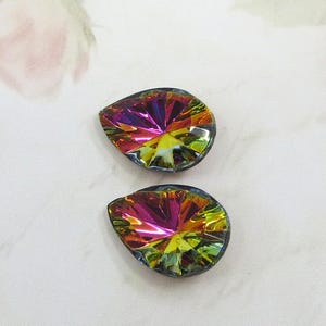 May include: Two iridescent teardrop-shaped crystal pendants. The crystals have a faceted design that reflects a spectrum of colors, including pink, purple, yellow, and green. The edges are outlined in a dark color, and the background is a soft, off-white.