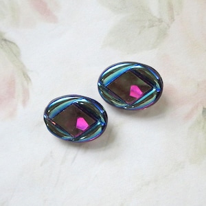 Op de afbeelding: Twee iriserende ovale knopen. Elke knoop heeft een centraal ruitvormig ontwerp in roze en paars, omgeven door een blauwgroene rand. De knopen liggen op een zachte, bloemige achtergrond.